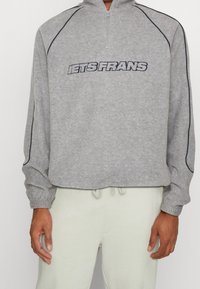 Grå sweatshirt med dragkedja, med texten "iets Frans" i svart och marinblå accenter längs axlar och ärmar.