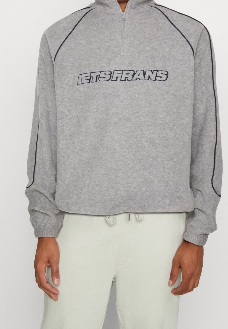 Grå sweatshirt med dragkedja, med texten "iets Frans" i svart och marinblå accenter längs axlar och ärmar.