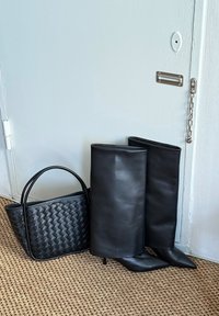 Bottes en cuir noir montantes avec embouts pointus à côté d'un sac à main tissé noir. Texture lisse et design structuré.