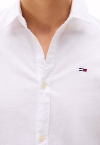 Camicia bianca button-up in lino, con colletto classico, bottoni gialli e un piccolo logo con accenti rossi, bianchi e blu sul petto.