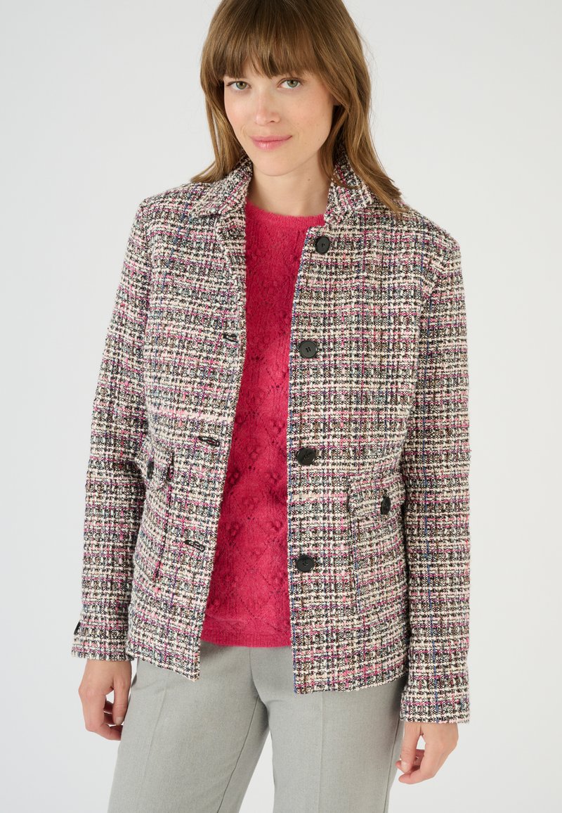 Giacca in tweed intrecciato rosa, nera e bianca con colletto, bottoni frontali e due tasche. Abbinata a un maglione rosa brillante.