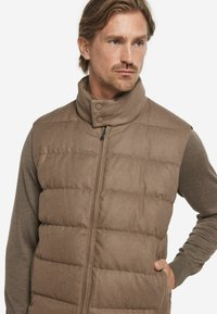 Braune Steppweste mit hohem Kragen, Frontreißverschluss und Druckknöpfen. Verfügt über eine gesteppte Struktur und Seitentaschen. Wird über einem Langarmshirt getragen.