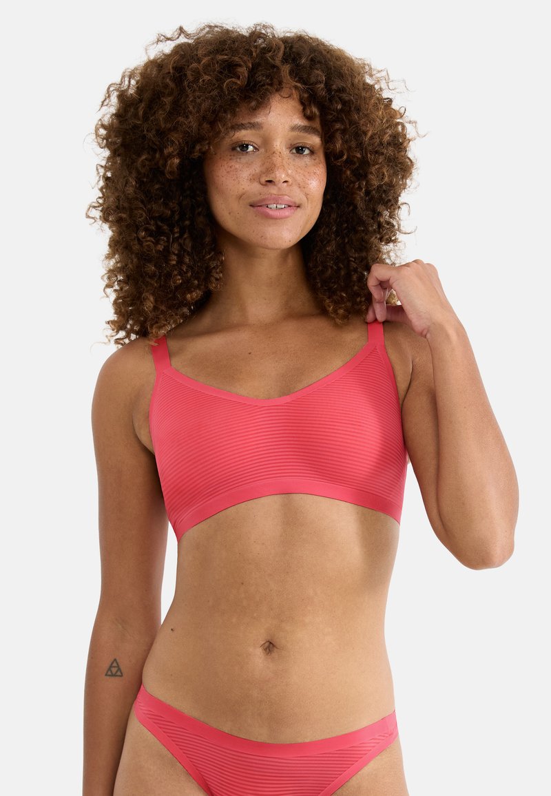 Frau mit lockigem Haar und Sommersprossen, die einen passenden korallenroten gerippten Bralette und Bikinihose trägt und mit einer Hand den Träger anpasst.