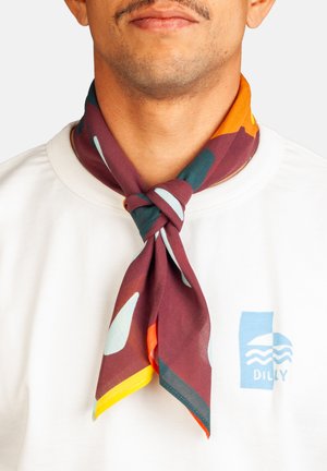 Hombre vestido con una camisa blanca con un logo azul, luciendo una bufanda de colores con un patrón atada alrededor de su cuello.