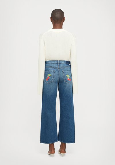 Persoon in hooggeknipte blauwe jeans met kersenborduursel op de achterzakken, in combinatie met een witte trui met lange mouwen en witte hakken, met de rug naar de kijker gekeerd.