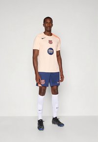Lys beige Nike fotballdrakt med oransje aksenter, Barcelona-logo og Spotify-grafikk. Kombinert med marineblå shorts og hvite sokker. Svarte fotballsko.