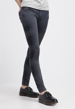 Personne portant un jean slim foncé, des chaussures noires à lacets et une chemise blanche avec des rayures horizontales noires, debout avec une main dans la poche.