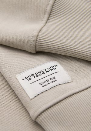 Beiger, gerippter Stoff mit einem eingenähten weißen Etikett, auf dem "Your only limit is your mind" und "OMBRE seit 1985" steht.