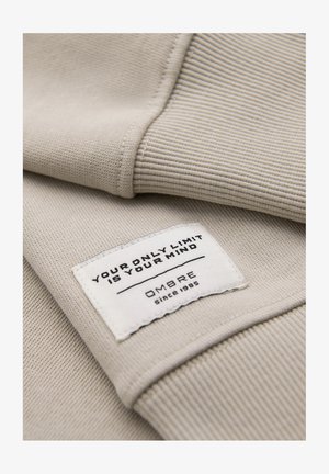 Beiger, gerippter Stoff mit einem eingenähten weißen Etikett, auf dem "Your only limit is your mind" und "OMBRE seit 1985" steht.