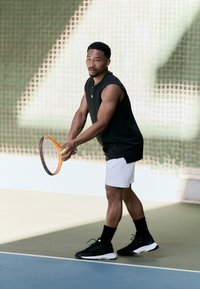 Juodas be rankovių sportinis marškinėlis ir balti šortai. Laiko oranžinį teniso raketę su geltonu kamuoliuku. Juodi sportbačiai ir kojinės. Teniso aikštelės fonas.