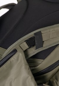 Gros plan d'une sangle rembourrée vert olive avec une boucle en plastique noir et des détails de couture sur un fond en tissu noir.
