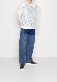 Felpa bianca con stampa di testo astratto in blu navy, abbinata a jeans in denim blu a vestibilità comoda e sneakers nere con suole bianche.