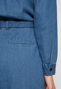 Gros plan d'une personne portant une robe en denim bleu avec une ceinture, une poche arrière et un poignet de manche longue avec un bouton noir.
