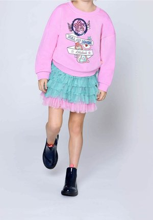 Enfant portant un sweat-shirt rose avec le motif "Pleine de magie", une jupe en tulle superposée turquoise et des bottines bleu foncé, marchant en avant sur un fond gris.