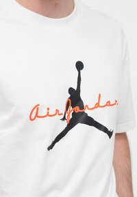 Άσπρο βαμβακερό μπλουζάκι με μαύρη σιλουέτα παίκτη μπάσκετ και πορτοκαλί κείμενο "Air Jordan". Κοντά μανίκια, στρογγυλή λαιμόκοψη.