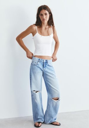 Jeune femme portant un débardeur blanc et un jean bleu clair déchiré et ample, ajustant la taille, debout contre un mur blanc.