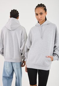 Felpa oversize grigia chiara con cappuccio regolabile, tasca a marsupio e polsini a costine. Il modello la indossa con leggings neri e jeans larghi.