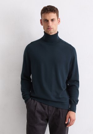 Mann in einem dunkelblauen Rollkragenpullover und dunkelgrauen Cordhosen, der vor einem schlichten weißen Hintergrund steht.
