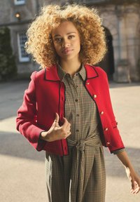 Nainen, jolla on kiharat vaaleat hiukset ja joka käyttää punaista takkia vyöllä varustetun ruudullisen mekon päällä, seisoo ulkona kivitalojen edessä.