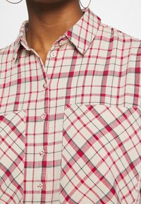 Gros plan d'une personne portant une chemise crème avec un motif à carreaux rouge, rose et noir, boutonnée, avec deux poches poitrine visibles.