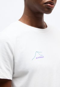 Κοντινό πλάνο ενός ατόμου που φοράει λευκό T-shirt με ένα μικρό μπλε περίγραμμα βουνού και τη λέξη "MAMMUT" σε μοβ στο στήθος.