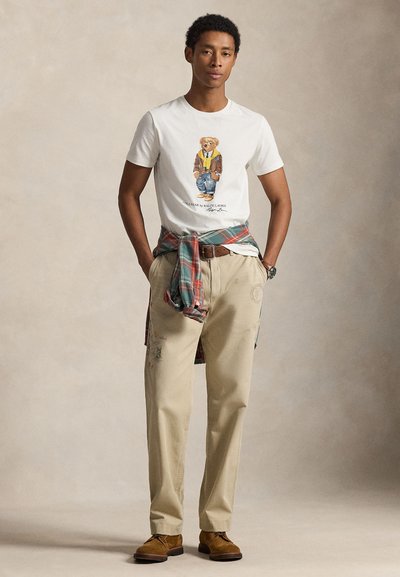 Tricou alb cu un desen cu urs, pantaloni chino bej cu detalii brodate și o cămașă în carouri legată în talie. Pantofi maro din piele întoarsă.