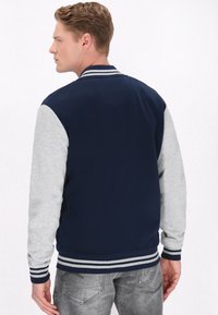 Mørkeblå varsityjakke med grå ærmer, ribbede manchetter og kant, samt stribe detaljer. Blødt stof, klassisk design, velegnet til afslappet brug.