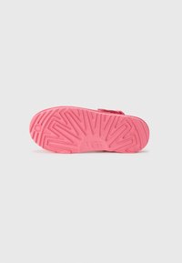 Rosa gummiskosula med ett strukturerat mönster som har spår och kanter, märkt med "UGG" i mitten. Ren, minimal design.