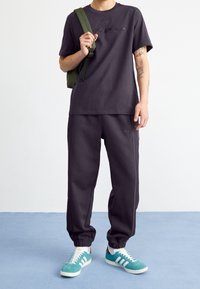 Camiseta de algodón gris oscuro con logotipo en relieve, combinada con pantalones de chándal a juego. Zapatillas deportivas verdes y blancas con tres franjas y detalles rojos.