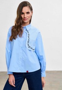 Femeie purtând o cămașă cu nasturi albastru deschis, cu un panou cu dungi și textul „Crește prin ceea ce treci”, combinată cu blugi albastru închis.