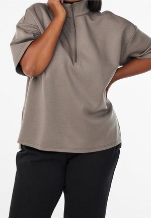 Persoon draagt een losse taupekleurige top met korte mouwen en een rits aan de voorkant, gecombineerd met een zwarte broek, poseert met één hand op de heup en de andere dicht bij het gezicht.