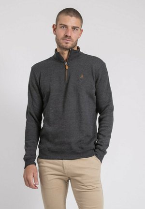 Homme portant un pull gris foncé à fermeture éclair quart, et un pantalon beige, debout avec une main dans la poche, devant un fond uni.