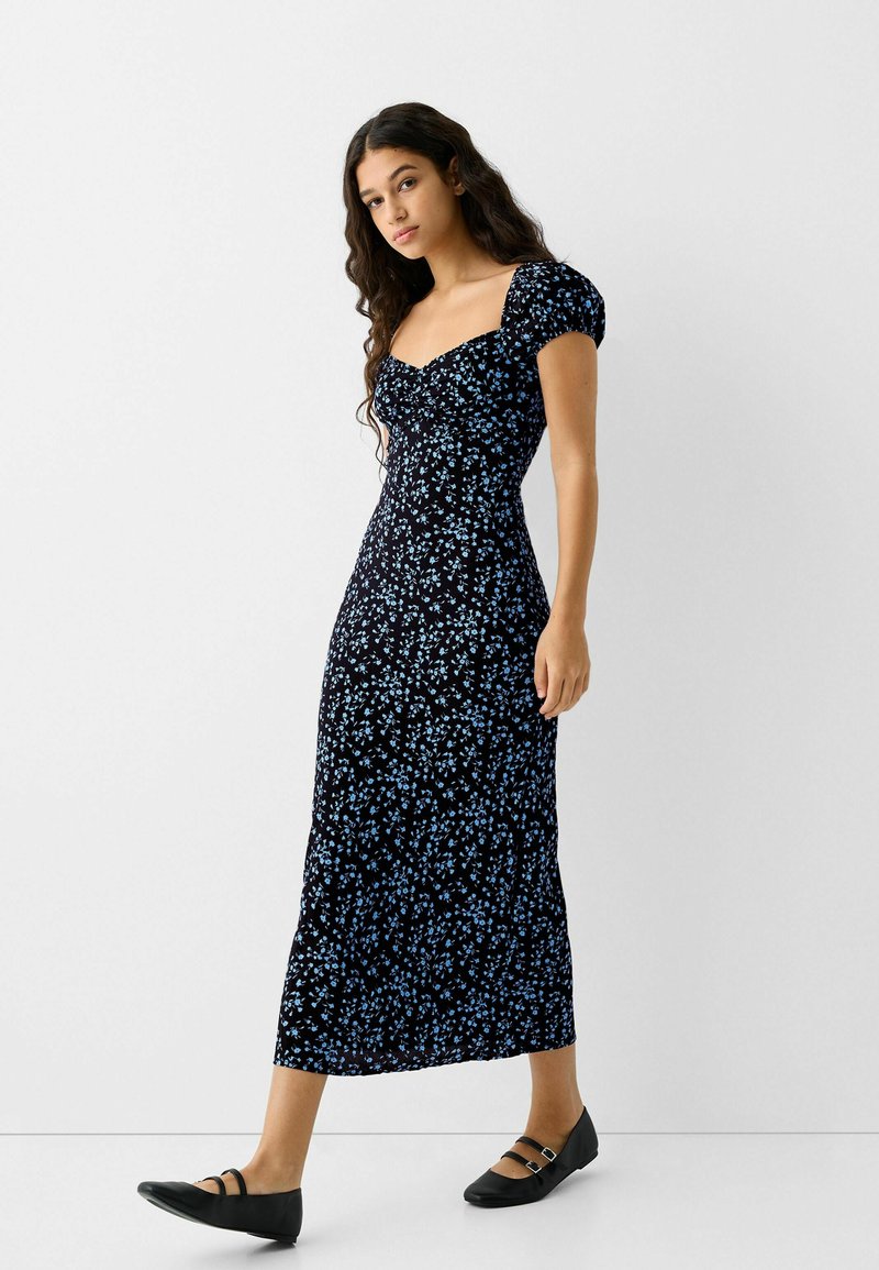 Vestido negro con flores que presenta flores azules, escote cuadrado, mangas cortas abullonadas y una silueta ajustada que llega hasta la mitad de la pantorrilla, combinado con bailarinas negras.