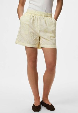 Femeie care poartă pantaloni scurți cu dungi galben deschis, talie elastică și buzunare, asortați cu pantofi negri comozi și un top alb.