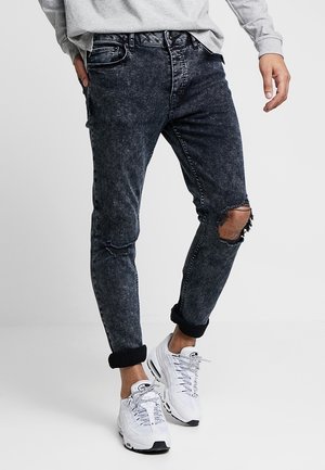 Mężczyzna ubrany w czarne, postrzępione jeansy z zakładkami, szarą bluzę i białe trampki, stojący na tle jednolitego, jasnego tła.