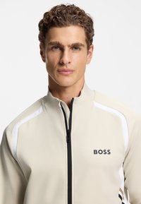 Beige zip-up jacket with white and brown accents, textured fabric, and "BOSS" logo on the left side. High collar and fitted design.