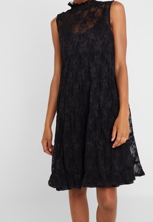 Robe noire sans manches en dentelle avec jupe à volants, col montant et corsage ajusté portée par une personne debout, les bras détendus.