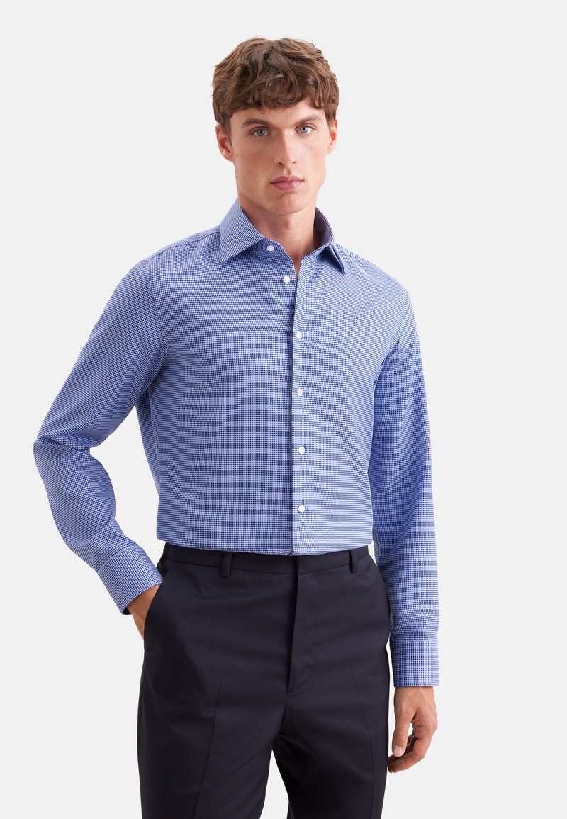 Jeune homme portant une chemise bleue à carreaux à manches longues rentrée dans un pantalon habillé noir, debout avec une main dans la poche sur fond blanc.