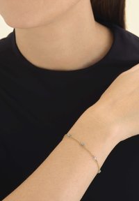 Zlatna narukvica s malim plavim kamenčićima ravnomjerno raspoređenima duž cijele dužine. Glatka tekstura na koži, minimalistički dizajn.