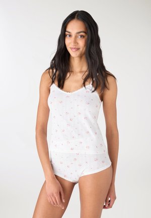 Giovane donna con lunghi capelli scuri indossa un top bianco con fiori e slip a vita alta abbinati, in piedi davanti a uno sfondo chiaro e uniforme.