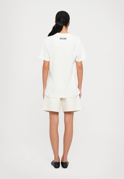 Tricou alb din bumbac cu mâneci scurte, având un mic logo negru "MOSCHINO" pe partea superioară a spatelui, asociat cu pantaloni scurți crema.