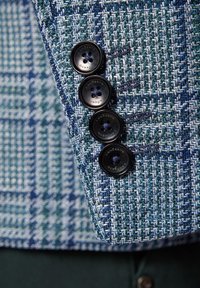 Niebieski i zielony żakiet w wzór houndstooth ma na rękawach cztery czarne guziki z kontrastującą nicią, prezentując strukturalną fakturę tkaniny.