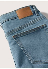 Jean en denim bleu clair plié montrant la poche arrière et l’écusson de marque en cuir marron à la ceinture sur fond blanc.