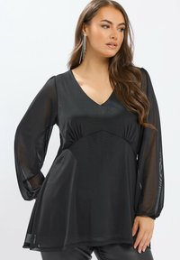 Tunique noire avec un col en V, des manches longues en translucide et un ourlet évasé. Présente une taille froncée et une texture lisse.