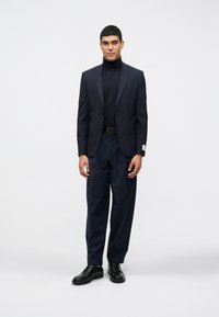 Homem vestindo um blazer azul marinho, uma blusa de gola alta preta, calças azul marinho com pregas, um cinto de couro preto e sapatos sociais pretos em pé contra um fundo branco.