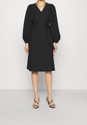 Femme portant une robe portefeuille noire texturée avec de longues manches bouffantes, une ceinture à la taille, un collier chaîne doré et des sandales à talons noires.