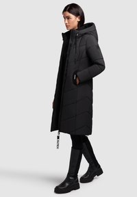 Manteau doudoune noir avec capuche, présentant un motif matelassé, une fermeture éclair, des poches latérales et une fermeture éclair de marque. Assorti à des bottes noires montantes.