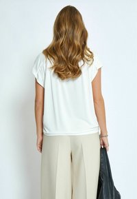 Top bianco a maniche corte con una vestibilità rilassata, abbinato a pantaloni larghi beige chiaro e una borsa nera in pelle in mano.