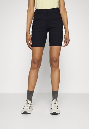 Person, die schwarze knielange Shorts, graue Socken und weiße Sneakers trägt, steht vor einem schlichten hellen Hintergrund.
