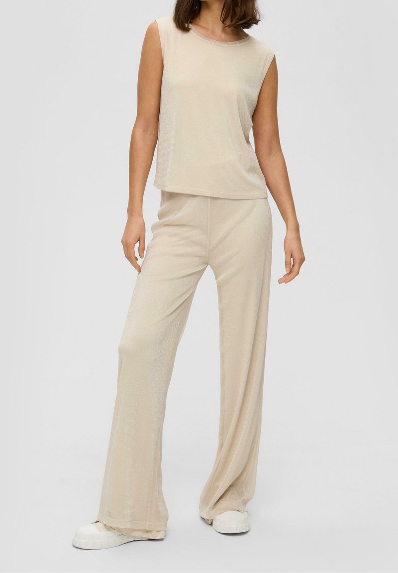 Femme portant un haut beige sans manches ample et un pantalon à jambes larges, associé à des baskets blanches, debout devant un fond uni.